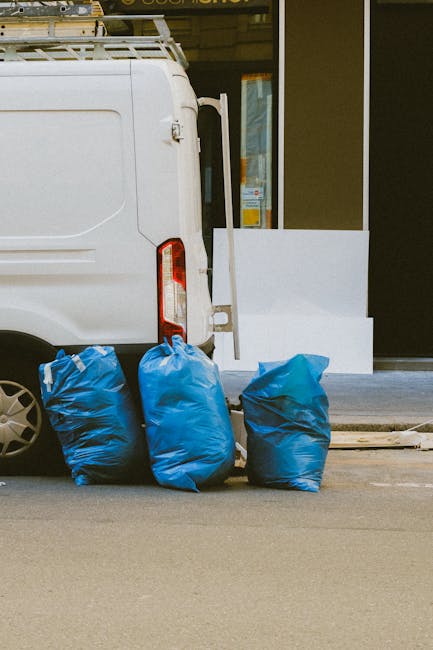 An open black van parked in a multi-story parking garage, filled with various cleaning tools and supplies including a metal shelving unit, cleaning bottles, and boxes. Outside the garage, there are gr