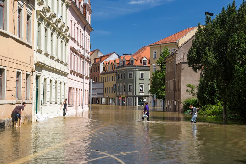 A silver hatchback car partially submerged in floodwater on a street, with people around it attempting to assist. A man is seen holding onto the rear of the vehicle, while others stand nearby on the f