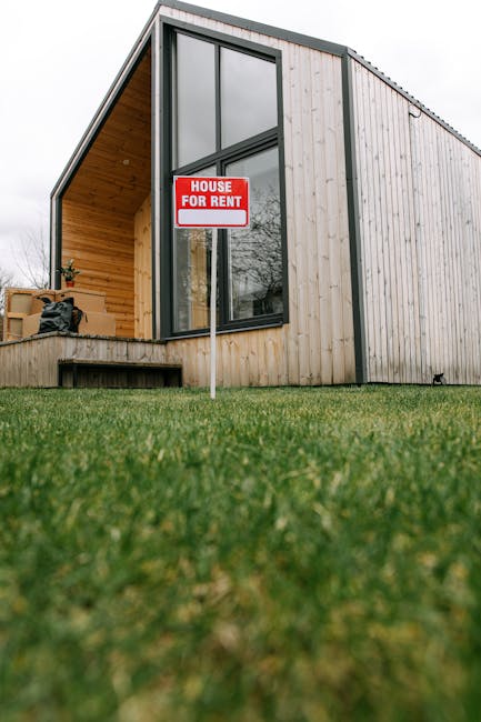 A red and white 'House for Rent' sign positioned on a grassy lawn in front of a modern building with vertical wooden paneling. The building features large black-framed windows and appears well-maintai