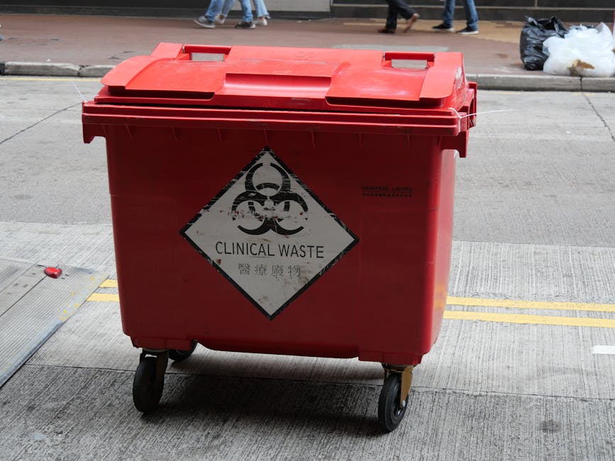 A person wearing a white protective suit, safety goggles, and a respirator mask is holding a yellow hazardous waste container while standing outdoors at a landfill site filled with scattered debris an